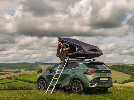 Man and dog sit inside the TentBox GO on top of a Kia Sportage, with rolling hills in the background.