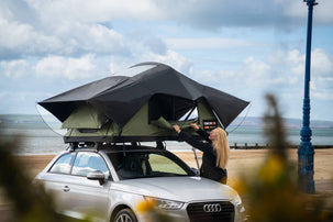 Woman opens door to Forest Green TentBox Lite 2.0, positioned on top of a silver Audi A1