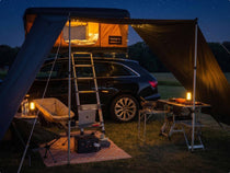 A campsite with a TentBox Classic mounted on the roof of a car, a tunnel awning set up and two TentBox Tripod Lights illuminating the area. 