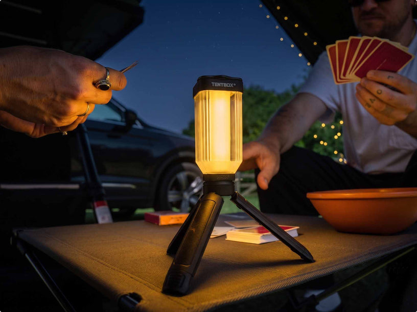 Lifestyle image showing the TentBox Tripod Light sat on a TentBox Camping Table, lighting up the area while two people play cards. 