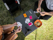 An overhead view of a TentBox Camping Table set on grass, with two people playing a card game. The table has the brand name 'TentBox' printed on it, along with playing cards, an orange bowl of snacks, and a smartwatch.