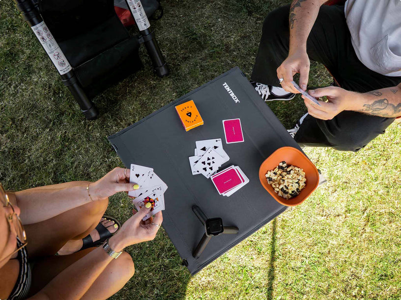 An overhead view of a TentBox Camping Table set on grass, with two people playing a card game. The table has the brand name 'TentBox' printed on it, along with playing cards, an orange bowl of snacks, and a smartwatch.