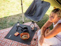 A woman sits beside a grey TentBox Camping Table set up under a tent outdoors. The table holds a bowl of snacks, a small bottle, and a can, with a grey TentBox Camping Chair and a patterned rug visible in the scene.