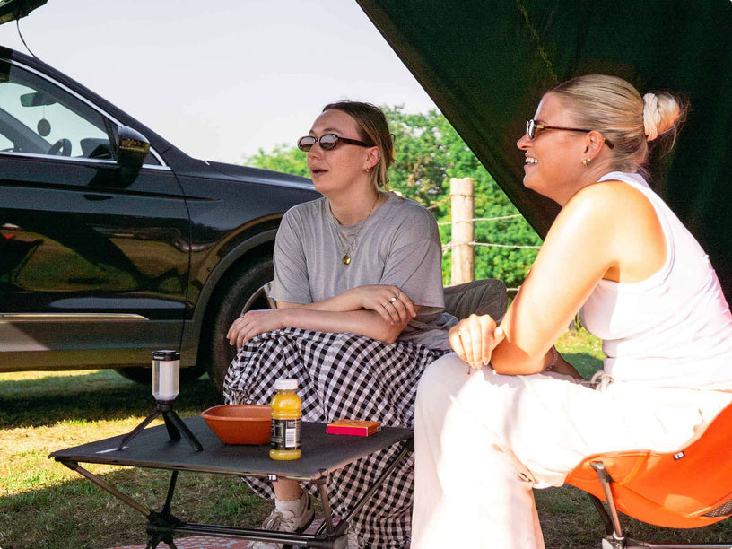 Two women sitting outdoors under an awning next to a black car, with a TentBox Camping Table holding a tumbler, bowl, and bottle between them.