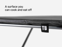 Close-up of a TentBox Camping Table on a metal frame, with a small black tag reading TB and the text 'A surface you can cook and eat off' on a light gray background.