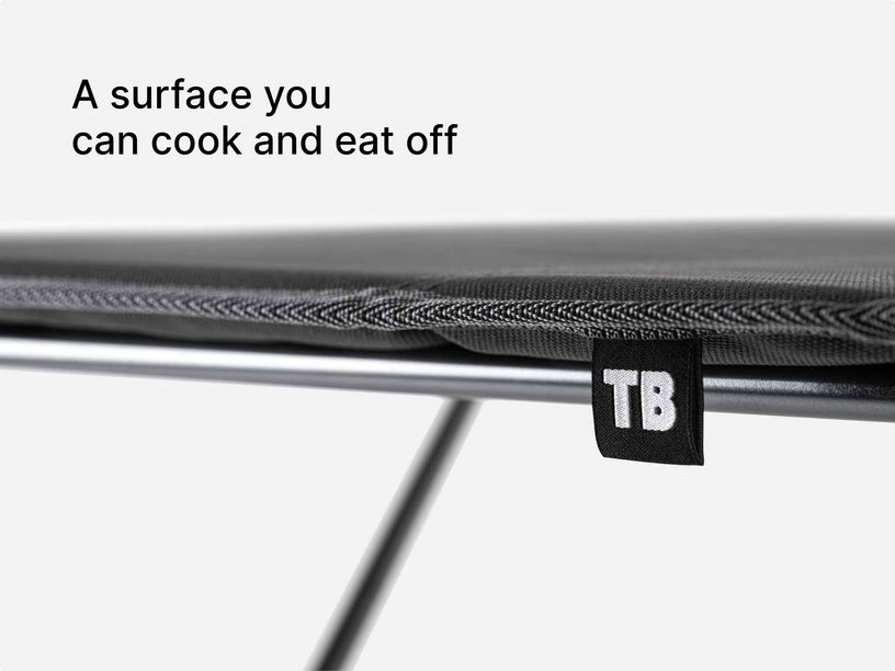 Close-up of a TentBox Camping Table on a metal frame, with a small black tag reading TB and the text 'A surface you can cook and eat off' on a light gray background.