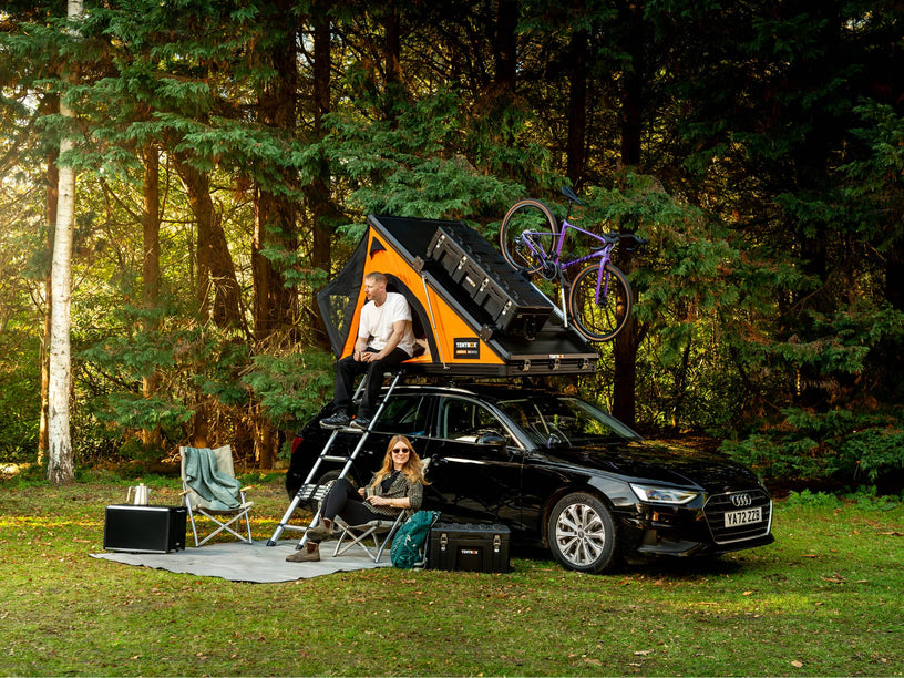 Two people camping in a forest with a car equipped with the TentBox Cargo 2.0 roof top tent and Cargo Bars