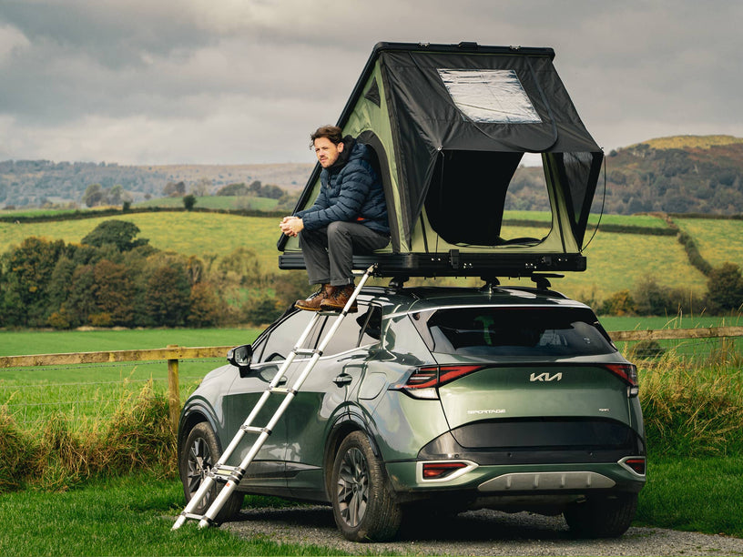 Person sitting on in the TentBox Cargo 2.0 roof top tent attached to a Kia SUV in a scenic outdoor setting.
