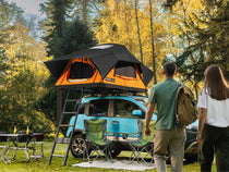 Two people walking towards a blue car with a new TentBox Lite roof tent in a campsite setting.