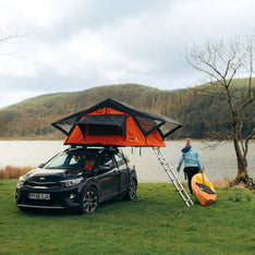 A woman carrying a canoe next to her TentBox Lite 1.0 Orange Edition which is mounted to a small hatchback car