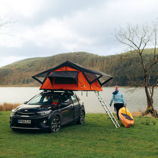 A woman carrying a canoe next to her TentBox Lite 1.0 Orange Edition which is mounted to a small hatchback car