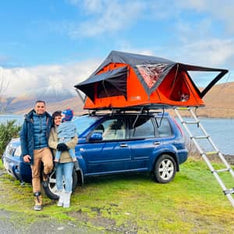 A young family smiling in front of their TentBox Lite roof tent, which is mounted to the roof of their family SUV car
